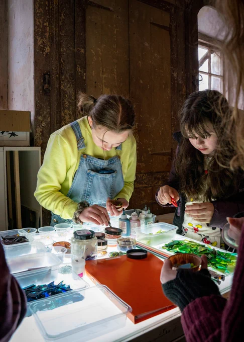Two young people stand in a dark room sorting pieces of coloured glass with tweezers. They look very focused.