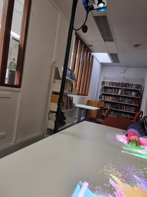 Image shows a white table in a library room which has colour splashes on it made by  a free standing projector above it. Just also see someone's hand moving across the projection to burst a colour balloon.