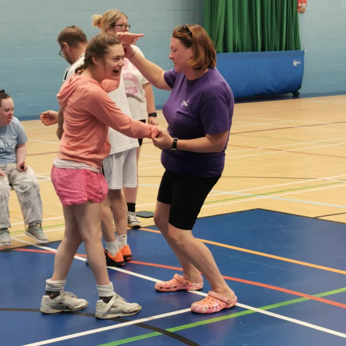 Izzie, a young woman with dark hair, dances in the middle of a sports hall on blue flooring. She is wearing a peach hoodie and pink shorts. The lady is assisted by a staff member wearing a purple Sense t-shirt. They are holding hands and moving together. There are others around them sitting and standing. 