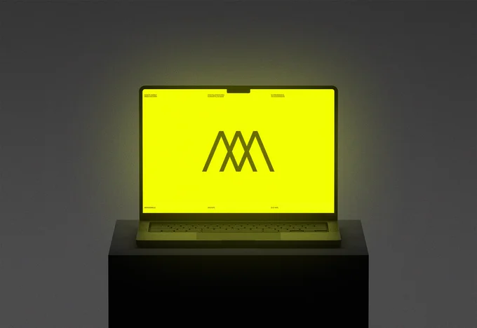 A laptop sitting on a dark cube. The laptop shows a yellow screen with a dark Monogram "M" logo in the center.