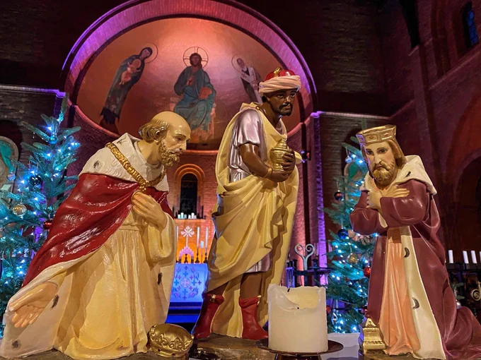 The foreground is a small candle surrounded by three large ceramic figures depicting the three Magi, or wise men. The background is a red brick church interior with coloured wall lighting and lit Christmas trees.