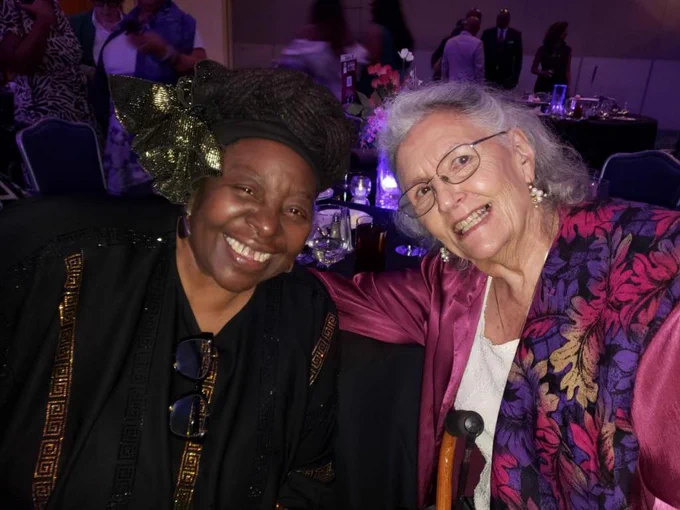An image of Loretta Ross and Norma Swenson: Norma is wearing a pink dress and ross is wearing a black and gold dress with a gold headband
