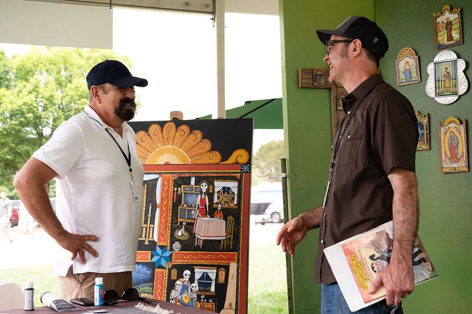 Two men laugh under a tent outdoors, surrounded by colorful paintings of saints.