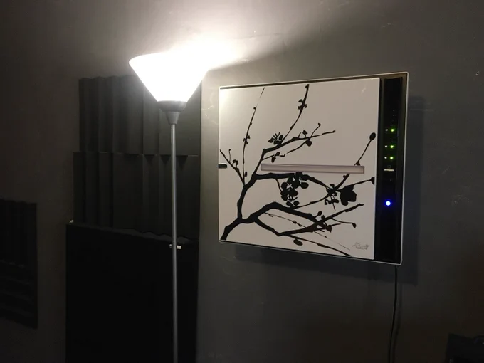 A MinusA2 air purifier mounted on a wall, featuring the 'Branch Out Zen' design on its panel, is illuminated by a floor lamp in a room with acoustic foam panels.