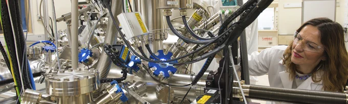 A scientist (a white woman in a lab coat and goggles) looking at a large, complex machine with many wires and metal pipes