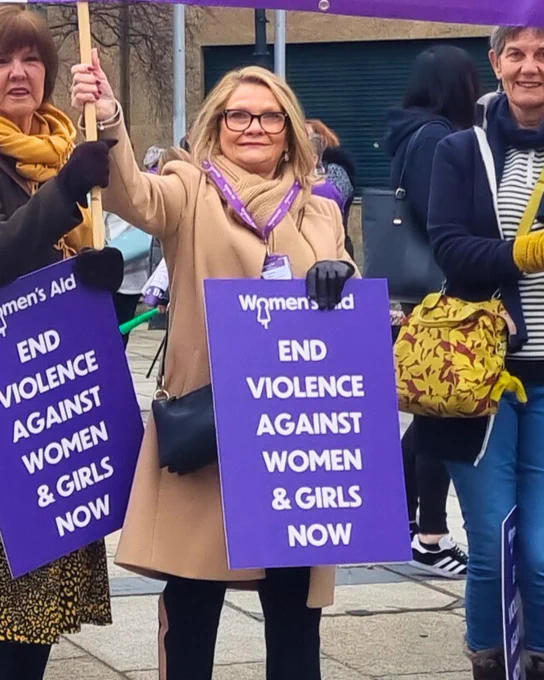 Image shows Sarah Mason, CEO of Women's Aid Federation Northern Ireland holding a sign saying "end violence against women and girls now" at the International Women's Day rally in Belfast in 2023.