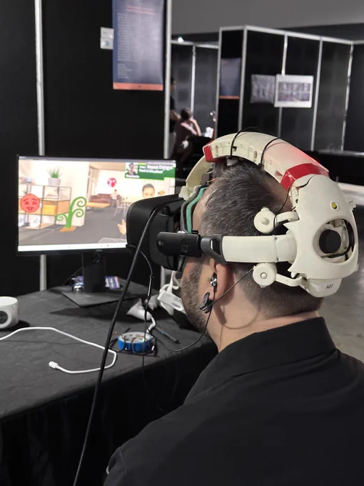 Participant at SIGGRAPH Asia experiencing the Healing Horizon VR demo using the Galea VR HMD. The individual is immersed in an adaptive virtual cafe scenario, with the headset's sensors visibly connected to monitor physiological responses. On-screen, the virtual environment depicts an interactive cafe setting.