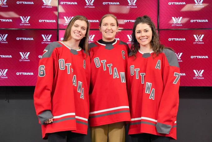 Emily Clark, Brianne Jenner and Jincy Roese posing for a photo wearing red PWHL Ottawa jerseys. Emily Clark and Jincy Roese are wearing an Alternate Captain’s “A” on their sweaters, and Brianne Jenner has the Captain’s “C”.