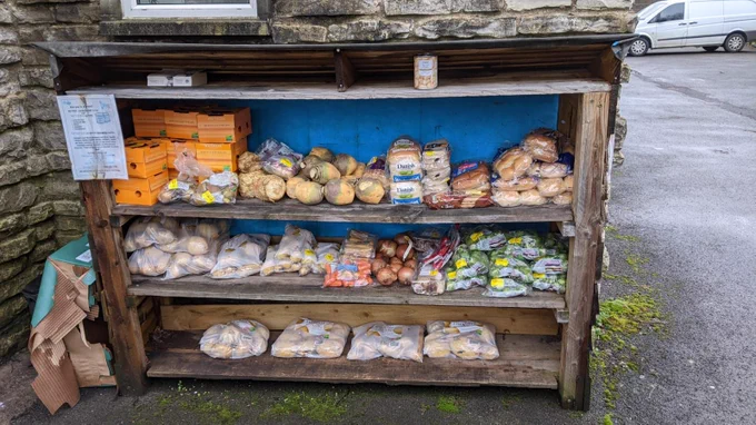 A community larder with wooden shelves full of donated surplus including oranges, swedes, bread, rolls, potatoes, carrots, parsnips, onions, mushrooms and brussels sprouts.