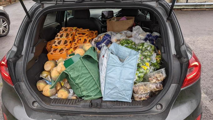 The boot of a car filled with various donated surplus food including oranges, brussels sprouts, swedes and potatoes.
