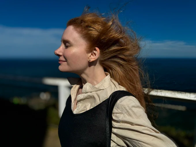 In a close-up profile, BDH stands with her eyes closed as the Australian summer breeze sweeps her hair back. She dresses in cream and navy linen and a slight smile grows on her face. In the background, the bright blue waters of Byron Bay.