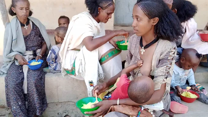 A mother breast feeding her baby while holding a bowl of food with several women and children sitting and eating behind her. 