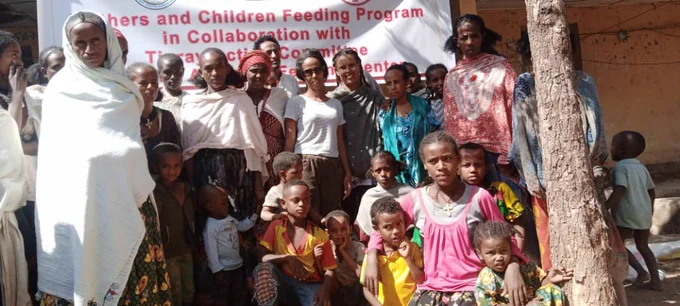 Women and children who are beneficiaries of Abergele feeding center stand and sit in front of the sign that reads ""Mothers and Children Feeding Program In Collaboration with Tigray Action Committee Yechilla Abergele Feeding Center". 
