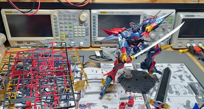 A fully assembled Dygenguar robot placed on top the assembly guide, with some electronics repair equipment in the background. The spent runners from the gunpla kit are next to it.