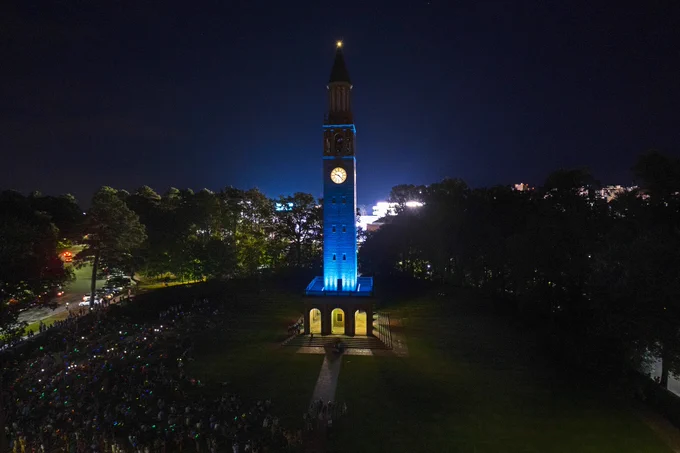 The UNC Morehead-Patterson Bell Tower lit up in Carolina Blue at night, surrounded by a dark sky and trees in silhouette.