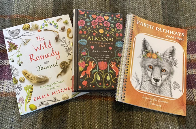 A photo of three books on a woollen blanket. The first is Wild Remedy Journsl, with a white cover and photos and sketches of nature: birds, shells, feathers, flowers. In the middle is The Almanac which has a black background with a folk art inspired brightly coloured design incorporating foxes, tulips, reminiscent of art nouveau in style. Then the Earth Pathways diary with an orange cover with a sketch of a Fox wearing a crown of the wild flower Fox and Cubs.