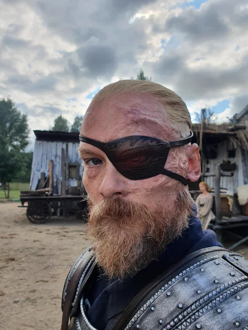 Close up of Guy Roberts as Uno Nomesta, in full armor standing in front of a shack on a beautiful grey day, looking directly at camera with a deadpan no shit Sherlock look on his face. Magnificent eyepatch and right good eye staring right into your soul. Atuans Mill set. Season 2 The Wheel of Time.