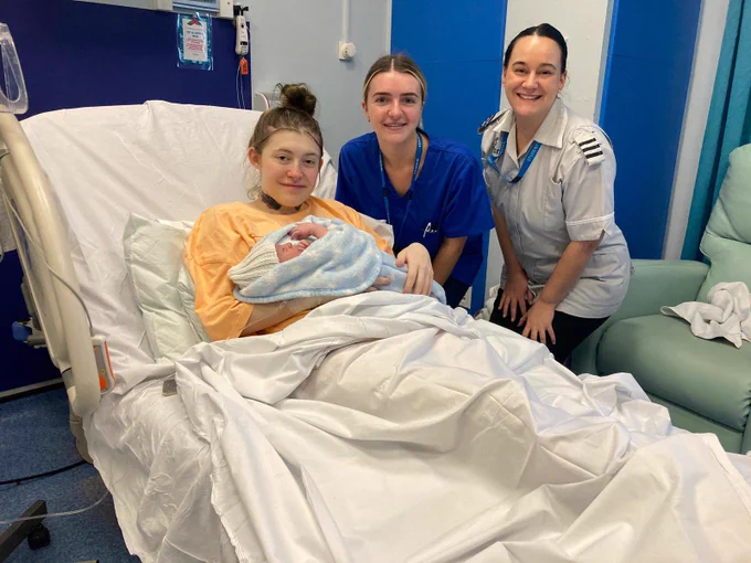 A young woman lays in a hospital bed holding a newborn baby with a midwife and student midwife next to her smiling