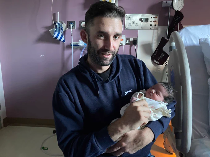 A man stands in a hospital side room holding a newborn baby