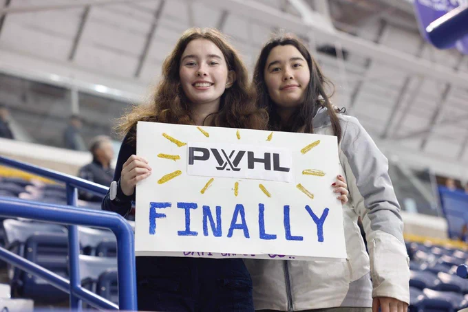 Two PWHL fans waiting for warm ups to start holding a homemade sign that reads "PWHL finally."