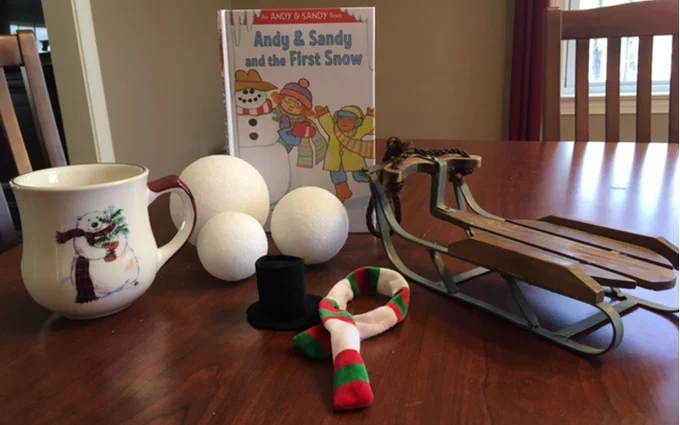 Pictured are items used in this lesson sitting on a brown  kitchen table. From left to right: a coffee mug with a polar bear on it,  three styrofoam snowballs in various sizes, the book “Andy & Sandy  and the First Snow,” a miniature top hat, a red, green and white striped  scarf and a wooden bobsled.