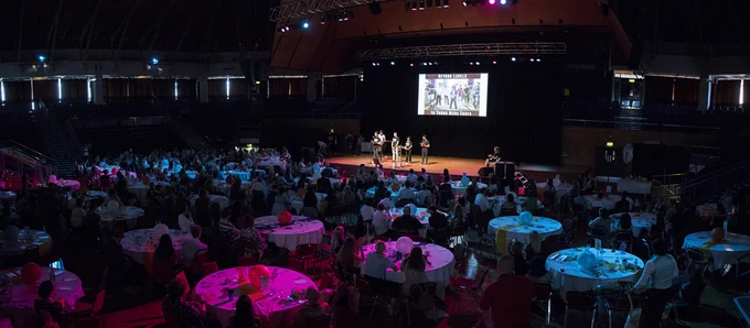 An awards ceremony room: people sat around round tables with a spoken word performance by young poets on stage in the distance. 