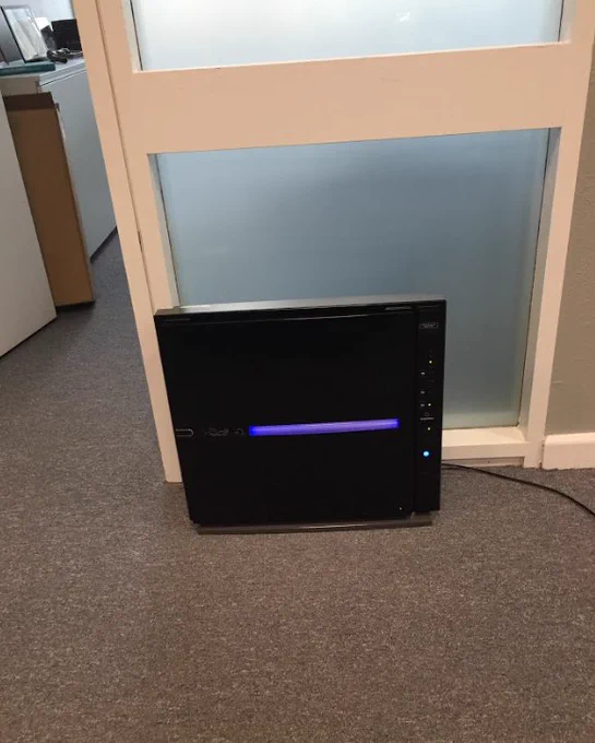 A sleek black MinusA2 air purifier with a vibrant blue mood light is situated on a gray carpet near a frosted glass door in an office setting.