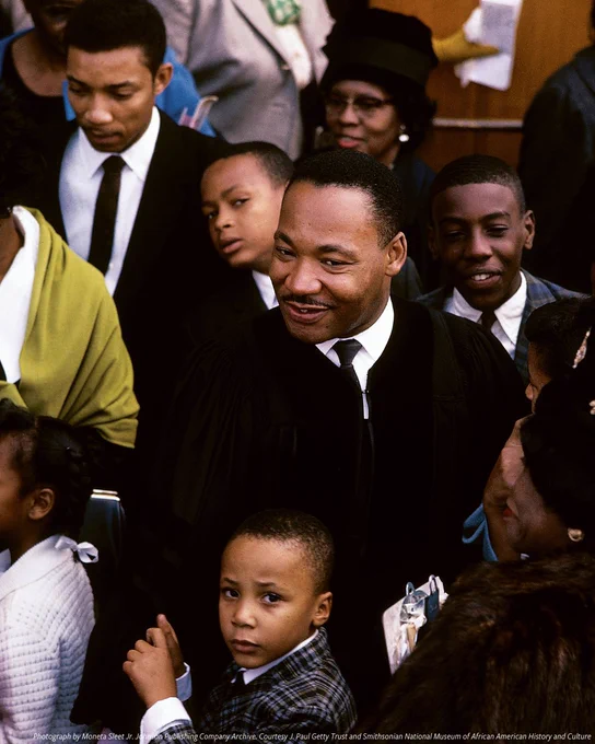  A color photograph of civil rights leader Dr. Martin Luther King Jr. standing in a crowd of mostly children as he receives a hug from his son, Martin Luther III. King's mother Alberta looks on from behind the group.