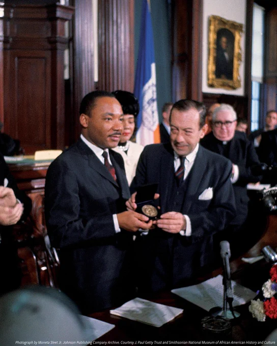 A color photograph from December 1964 of Dr. Martin Luther King Jr. standing next to a man in a suit and tie surrounded by a group of onlookers. Both hold up King's Nobel Peace Prize with both hands. His wife Coretta Scott King is partially visible behind the two men.