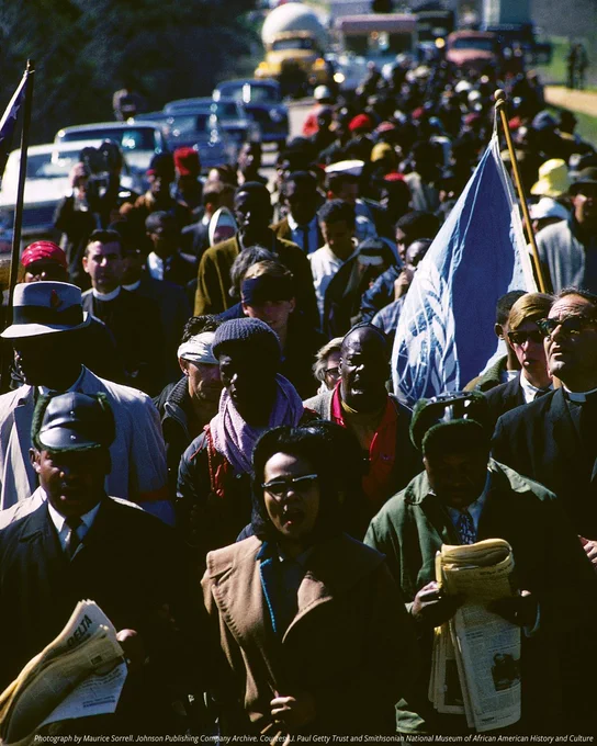 A color photograph of Dr. Martin Luther King Jr., his wife Coretta Scott King and Rev. Ralph Abernathy leading a group of  marchers toward Alabama's state capitol in Montgomery on March 21, 1965. A row of cars and trucks are visible on to the left of the marchers, and a marcher a few rows behind the Kings holds up a large blue flag. 