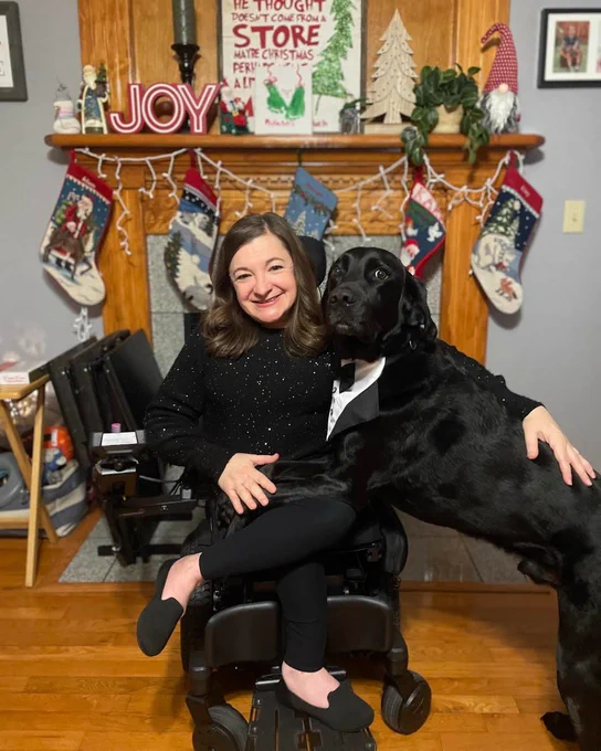 Photo of a woman and her dog in front of a mantle that is decorated for the holidays. She is wearing a black sweater and her pup is wearing a tuxedo bib collar.