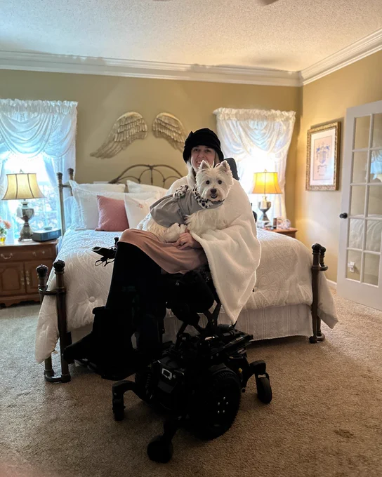 Photo of a woman with her dog in a bedroom. She is wearing a hat and a pink dress. The pup is a white terrier wearing a gray sweater.