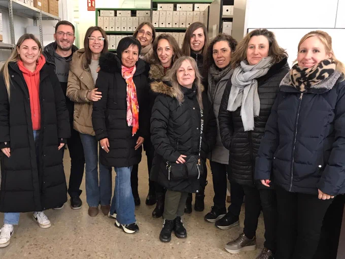 Fotografia del grup de dones del programa de formació de l'Institut Municipal d'Ocupació Treball i Formació (Línia DONA) al dipòsit de Gardeny de l'Arxiu Municipal de Lleida. El grup està format per un total de nou dones més la professora. A la fotografia també apareix el tècnic de l'Arxiu que va fer la visita i es poden veure al fons les prestatgeries i els armaris compactes amb capses d'arxiu. 