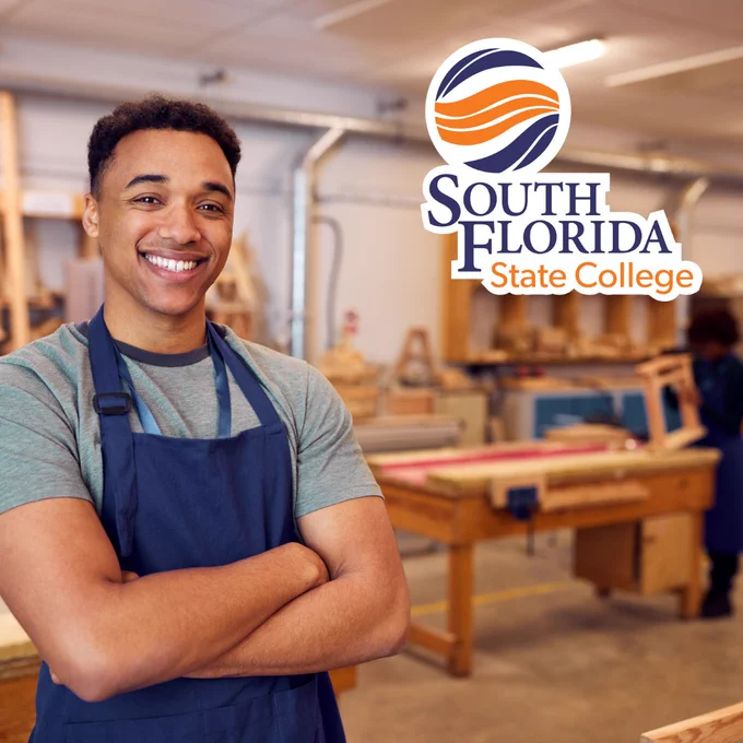 A student in a blue apron standing in front of a wood table while someone in the background builds an item out of wood. 