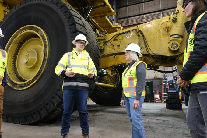 Senator Murray is standing next to a man, both of them are wearing hard hats and safety vests. Behind them is a large yellow truck.