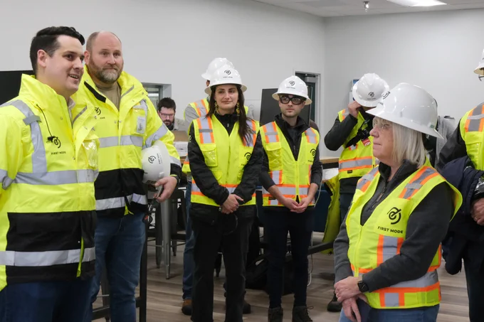 Senator Murray is standing on the right wearing a white hard hat and a safety vest. There are several people around her, all looking on and the same safety equipment. They're standing in a room with white walls.