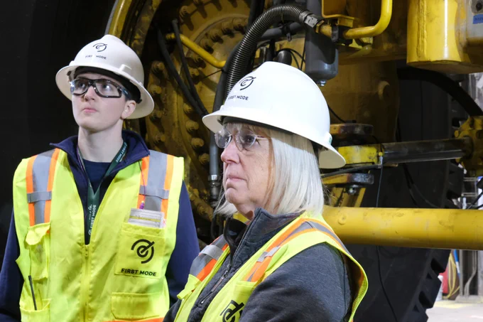 Senator Murray is in the foreground on the right and looking to the left. She is wearing a white hard hat and safety vest and has a serious expression.  Behind her is a man who is also in safety equipment. In the background there is part of a large yellow truck.