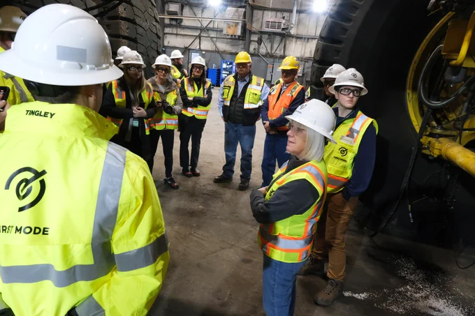 Senator Murray is on the right, standing in a white hard hat and safety vest looking to the right. There is a group of people in the same safety equipment standing around her in a shop.