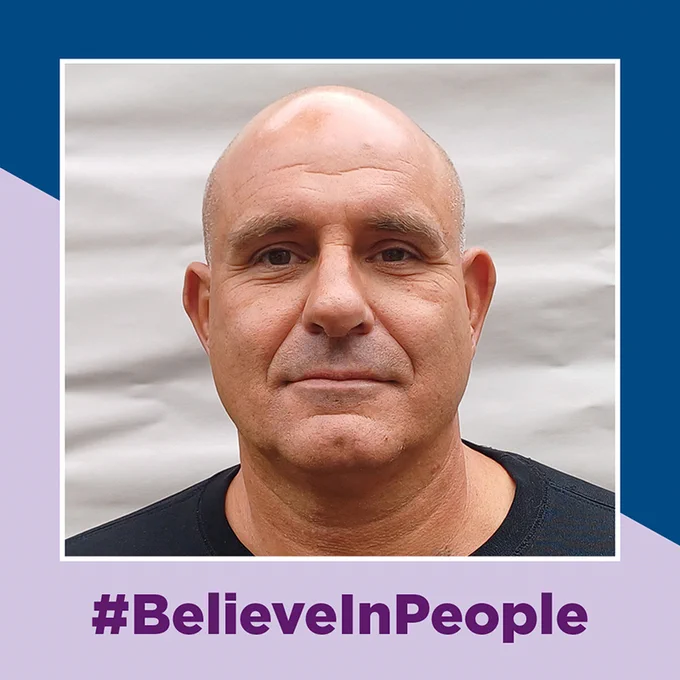 A blad man smiling at the camera. the photo sits on a background of dark blue and lilac with the words #BelieveInPeople underneath