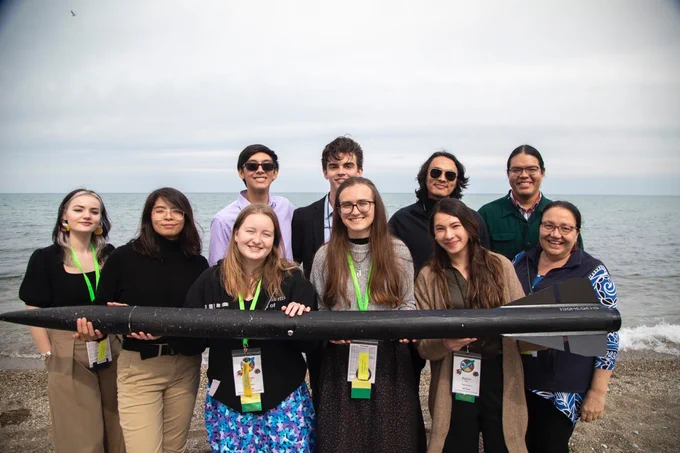 10 team members pose for a group photo on the beach while holding a long rocket in their hands.
