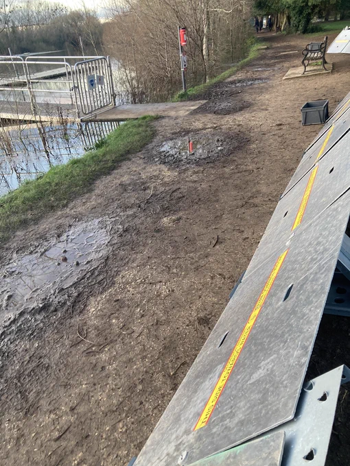 Metal barrier erected at edge of Hinksey Lake, with metal barrier on right hand side of picture and muddy looking bank of river and water on the left hand side.