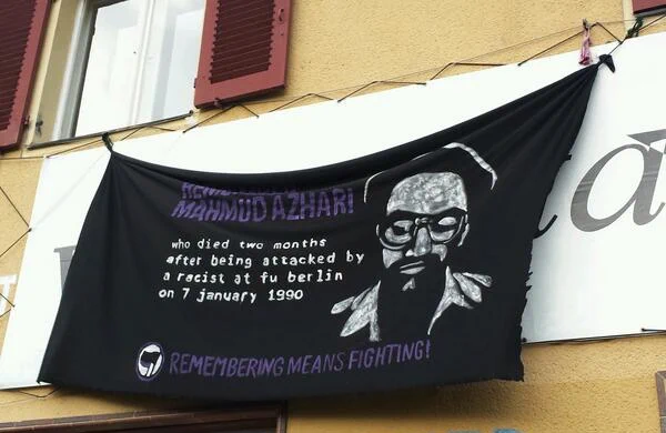 Schwarzes Transparent mit lila und weißer Schrift sowie einem Schablonenartigen Bild von Mahmud Azhar. Auf dem Transparent steht: "Remember Mahmud Azhar who died two months after being attacked by a racist at fu berlin an 7 january 1990 - Remembering means Fighting".
