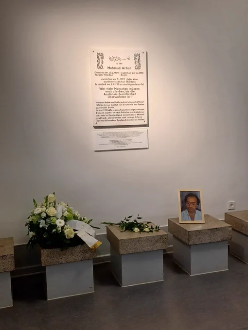 Gedenktafel für Mahmud Azhar an einer weißen Wand, direkt davor liegen ein Blumenkranu und ein kleiner Blumenstraße sowie ein gerahmtes Bild von Mahmud Azhar.