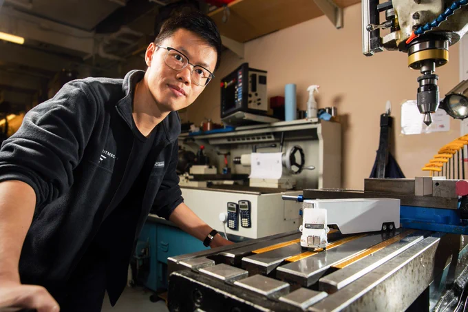 ZhiYi Liang, Asian man wearing a black t-shirt and dark jacket, clear glasses, short hair. Stands in a lab with a small model of a white truck.