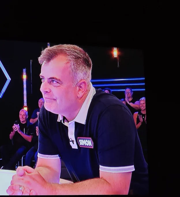 Simon Gregson leaning on the deal or no deal desk, smiling. 