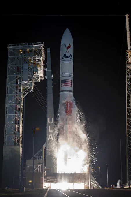A remote camera image of the first flight of the United Launch Alliance (ULA) Vulcan rocket as it lifts off its launch platform. The white rocket in the center of the image has the ULA logo and the American flag painted along its length, and sparks and fire emanate from its base. Its metal gantry to the left, and the road in the foreground are illuminated by the fire’s glow. Credit: United Launch Alliance