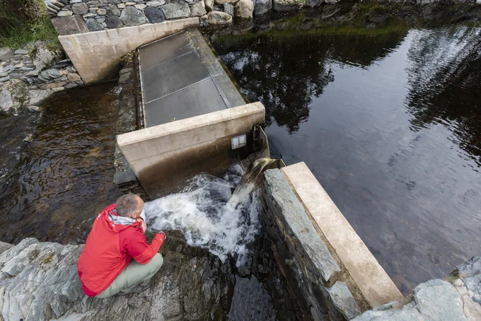 A National Trust ranger wearing a red rain jacket inspects a mountain hydro, a flat concrete structure which generates energy using water.