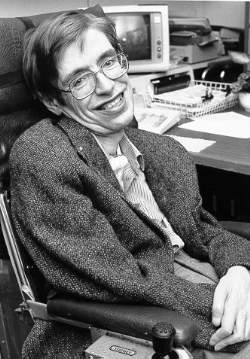 Picture of smiling man about 40 years old with glasses and arms crossed. He is in a wheel chair and a computer is behind him on his desk.