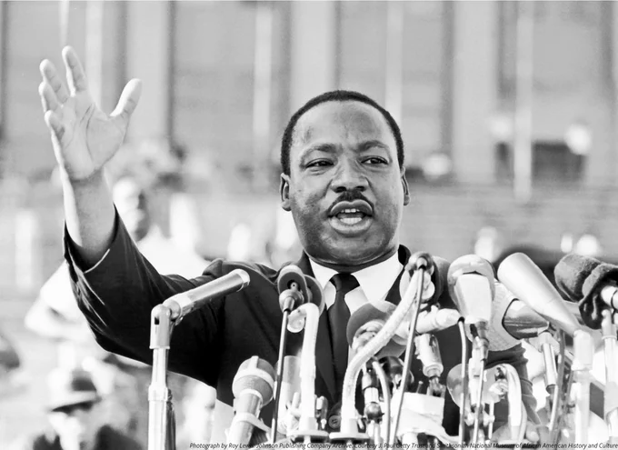 A black and white photo of Dr. Martin Luther King Jr. addressing a crowd at Chicago's Solider Field in 1966. He stands behind a podium and a choir of mounted microphones. 