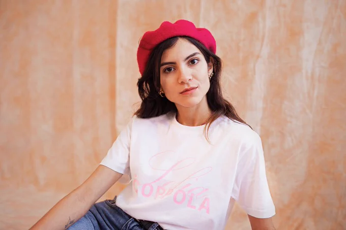 A young model wearing a red beret poses with the Sofia Coppola Priscilla special edition Girls on Tops tee in front of a peach background